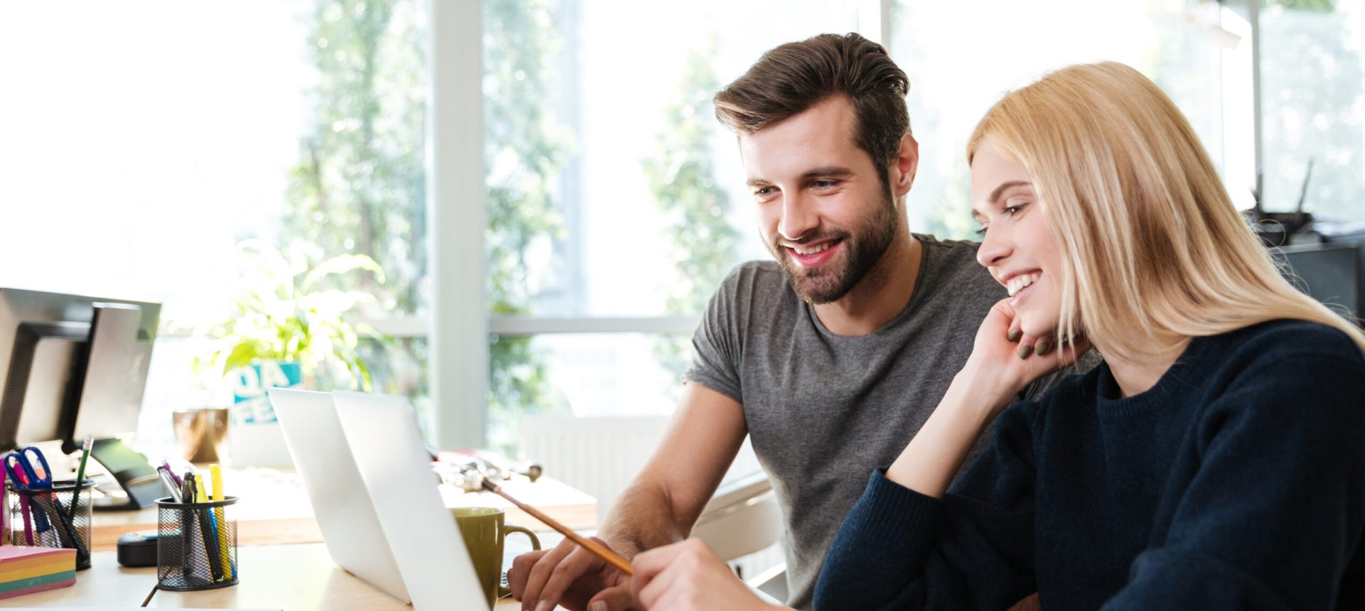 Happy young colleagues sitting in office coworking using laptop Image of happy young colleagues sitting in office coworking using laptop computers. Looking aside.